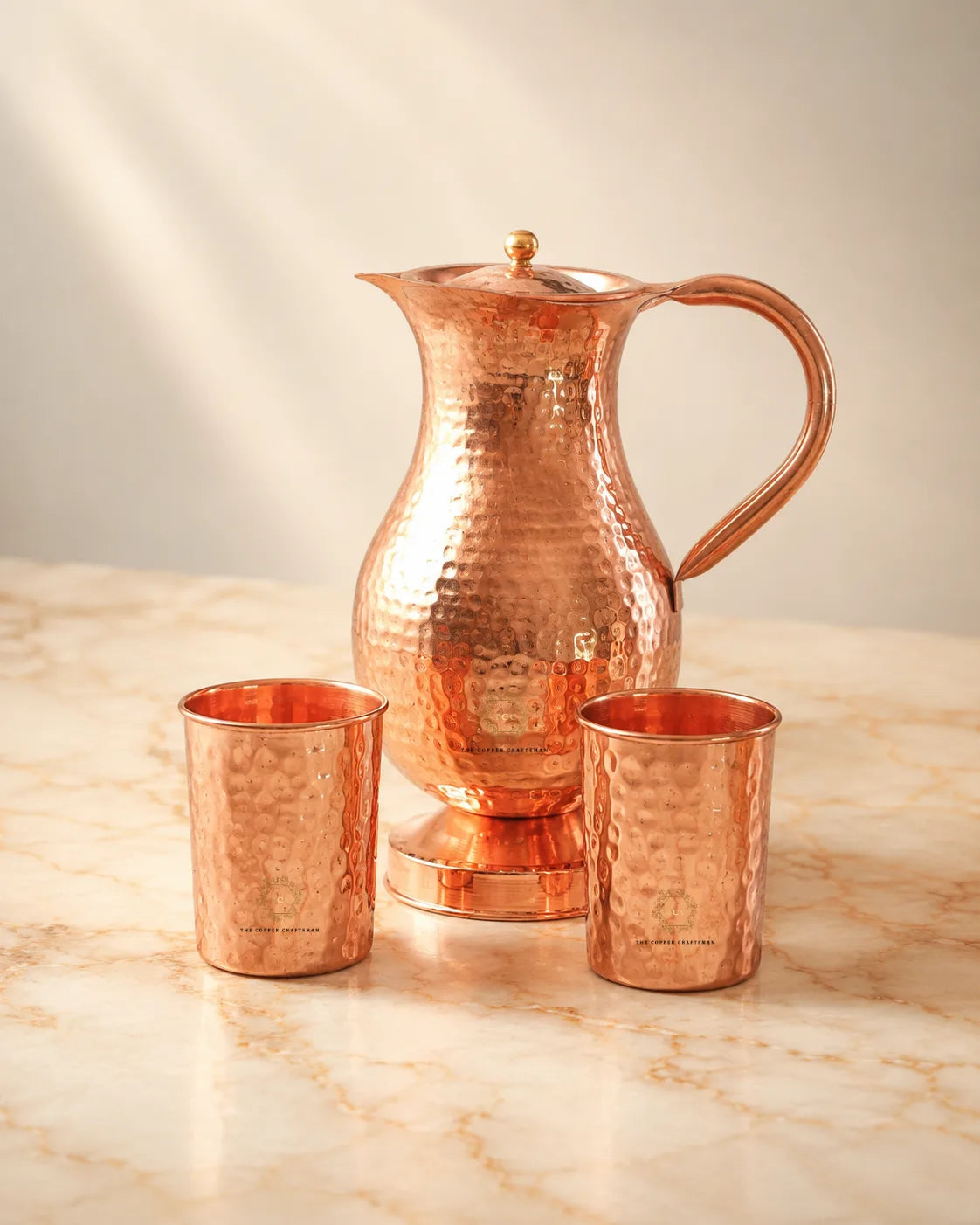 A hammered copper water pitcher with a matching lid and two small copper cups, arranged on a light marble surface against a soft beige background.
