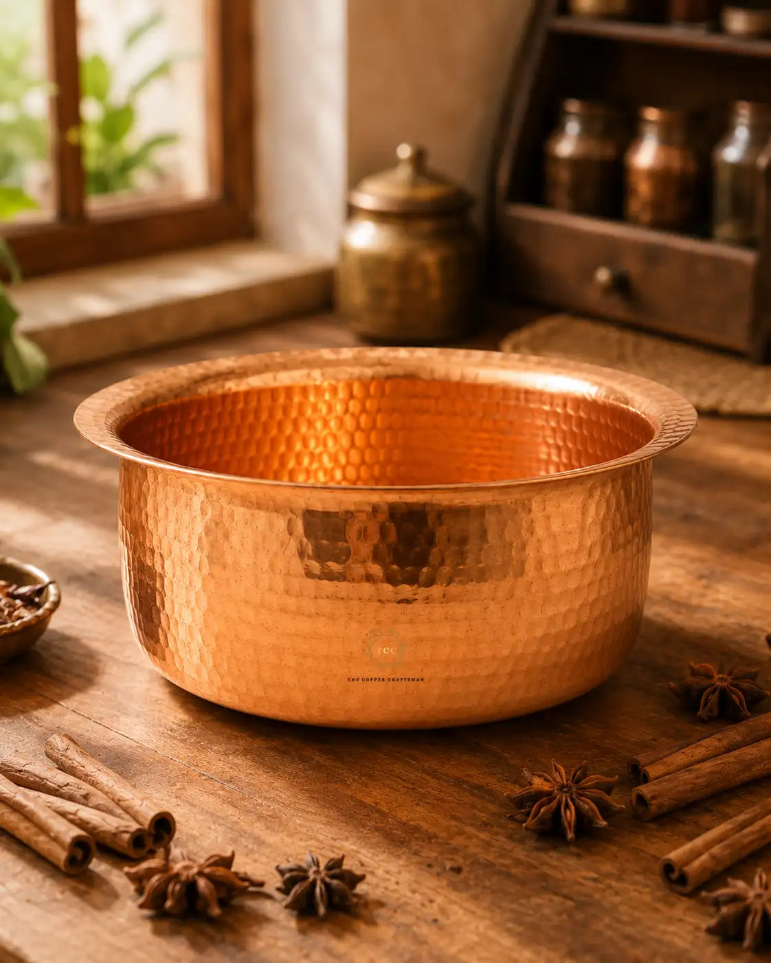 A Hammered Copper Cooking Pot (4.2 Litres) with a flared rim, displayed on a rustic wooden table surrounded by cinnamon sticks, star anise, and spices. In the background, a traditional brass jar sits on a windowsill with a wooden shelf holding glass bottles.