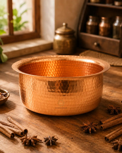 A Hammered Copper Cooking Pot (4.2 Litres) with a flared rim, displayed on a rustic wooden table surrounded by cinnamon sticks, star anise, and spices. In the background, a traditional brass jar sits on a windowsill with a wooden shelf holding glass bottles.