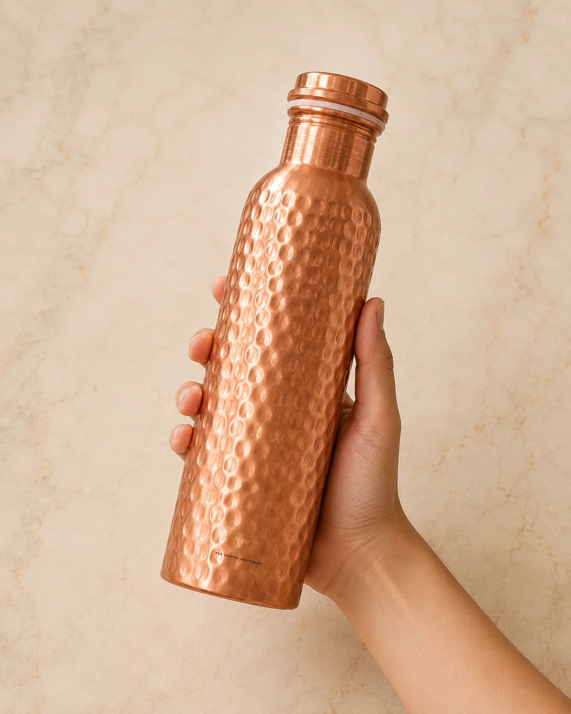 A hand holding a hammered copper water bottle against a light beige marble background.