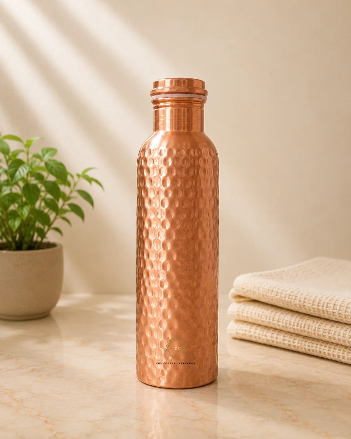 A hammered copper water bottle with a matching cap, standing on a marble countertop next to a potted plant and folded cream towels.