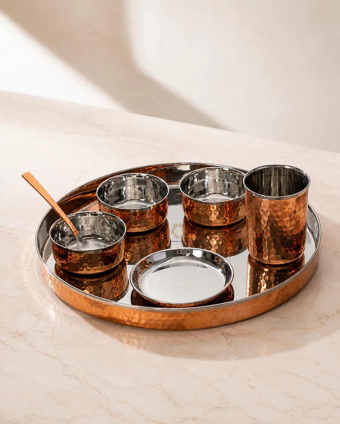 A Copper Dinner Thali Set with Stainless Steel Interior – 7 Piece  set arranged on a round hammered tray, placed on a light marble surface with a neutral background.