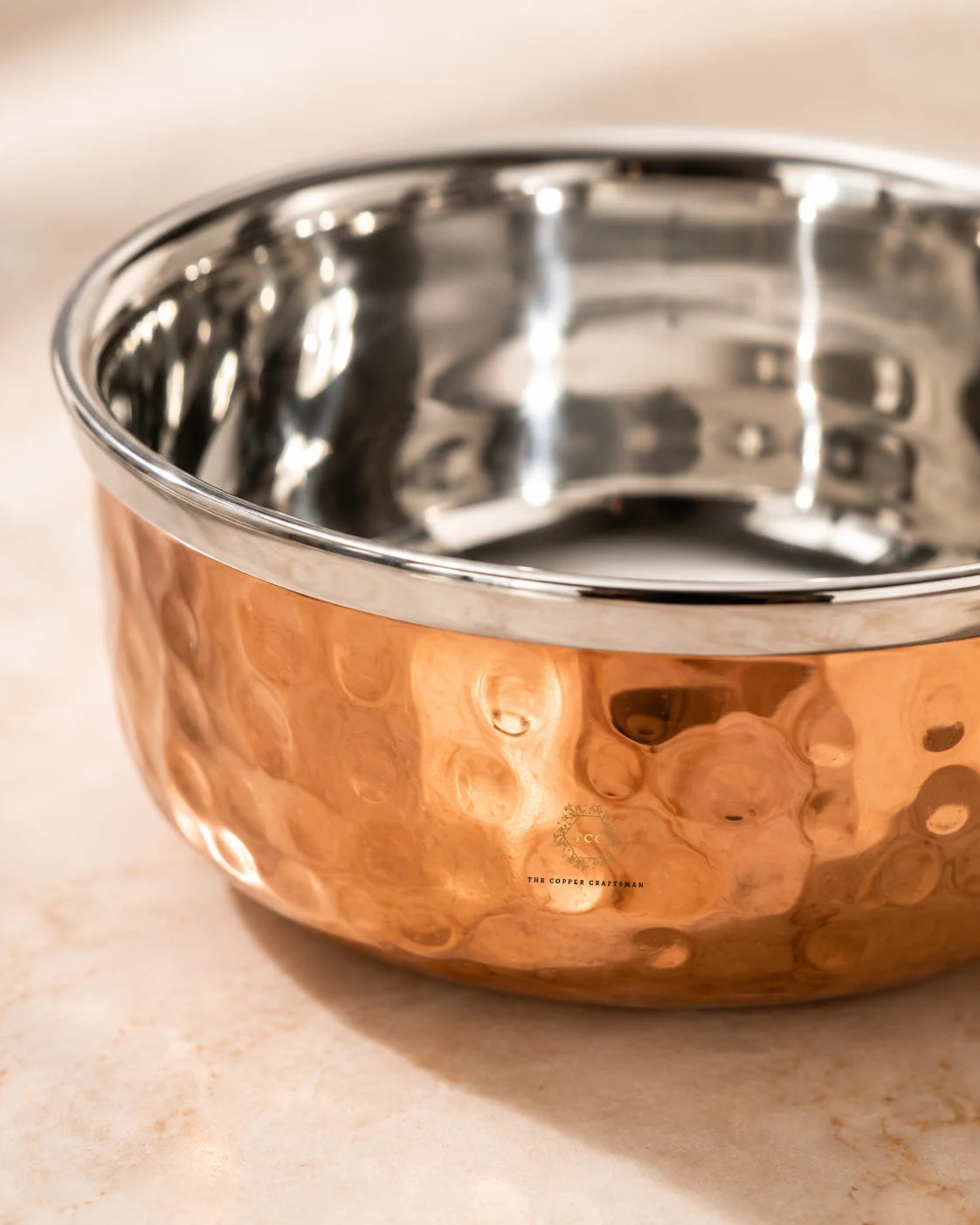 A hammered copper and interior polished stainless steel bowl shown in a close-up shot on a light beige marble surface.