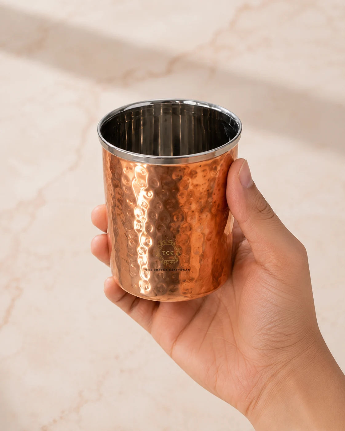 A hand holding a hammered copper tumbler with a polished stainless steel interior, set against a light beige marble background.