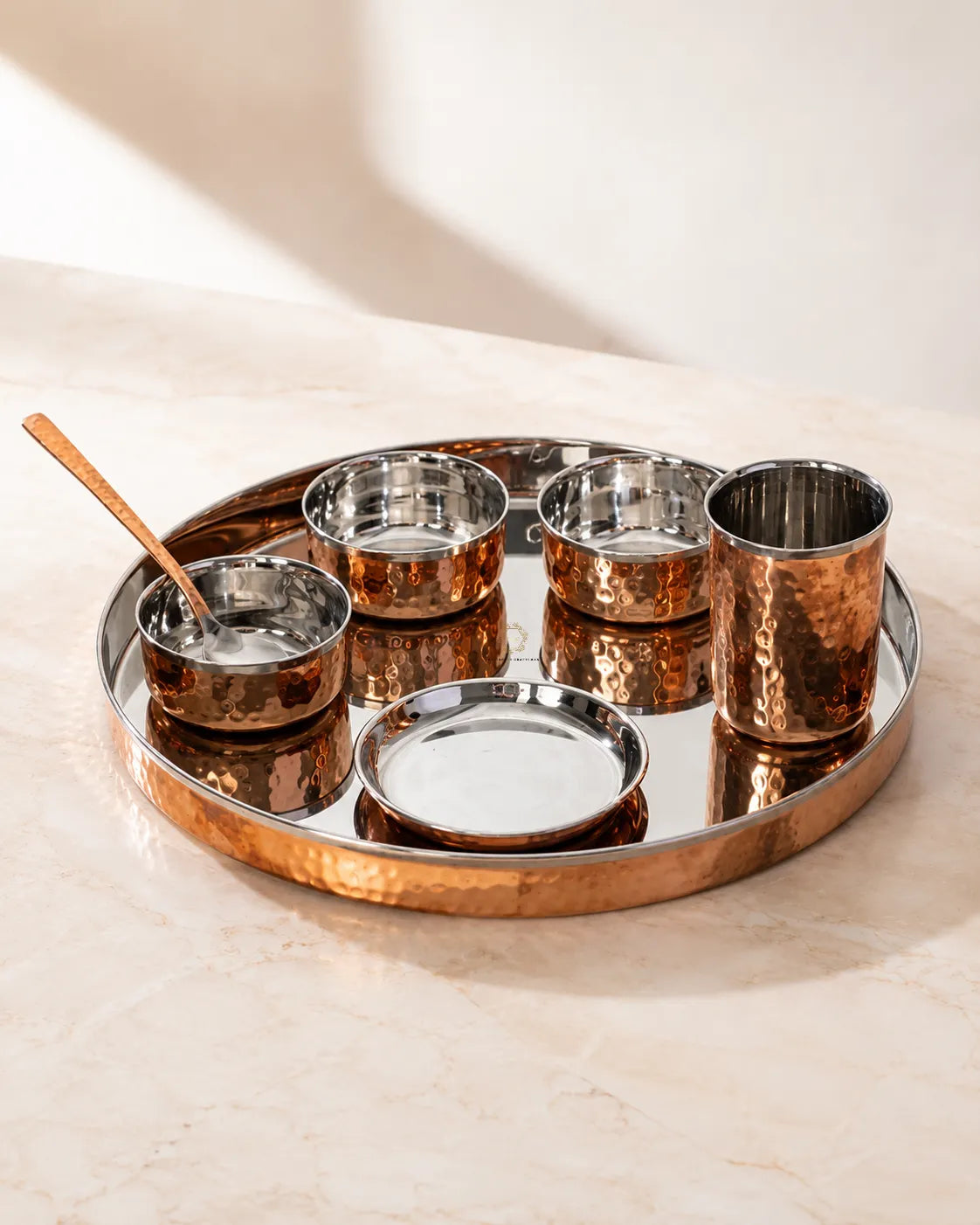 A Copper Dinner Thali Set with Stainless Steel Interior – 7 Piece set on a round tray, including four small metallic cups, a shallow dish, a spoon, and a lid, arranged on a light stone surface.