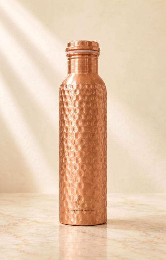 A hammered copper water bottle with a matching screw cap, standing upright on a light marble surface against a warm beige background with soft diagonal shadows.