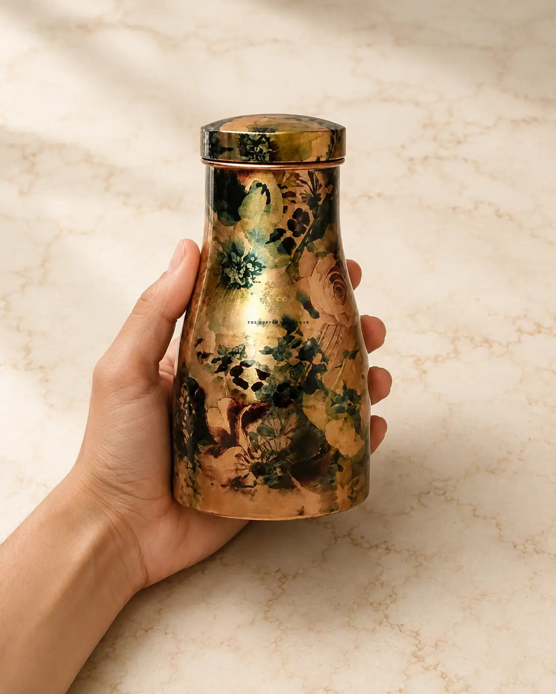 A hand holding a  pure copper water jar 1.5 litre with a metallic gold finish and a floral pattern in dark green, black, and warm brown tones, set against a light beige marble background.