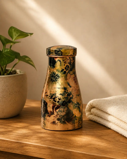 A Hand-painted floral pure copper water jar 1.5 litrewith a gold-toned finish and dark floral patterns, standing on a wooden surface in a softly lit interior setting with a plant and folded towel nearby.