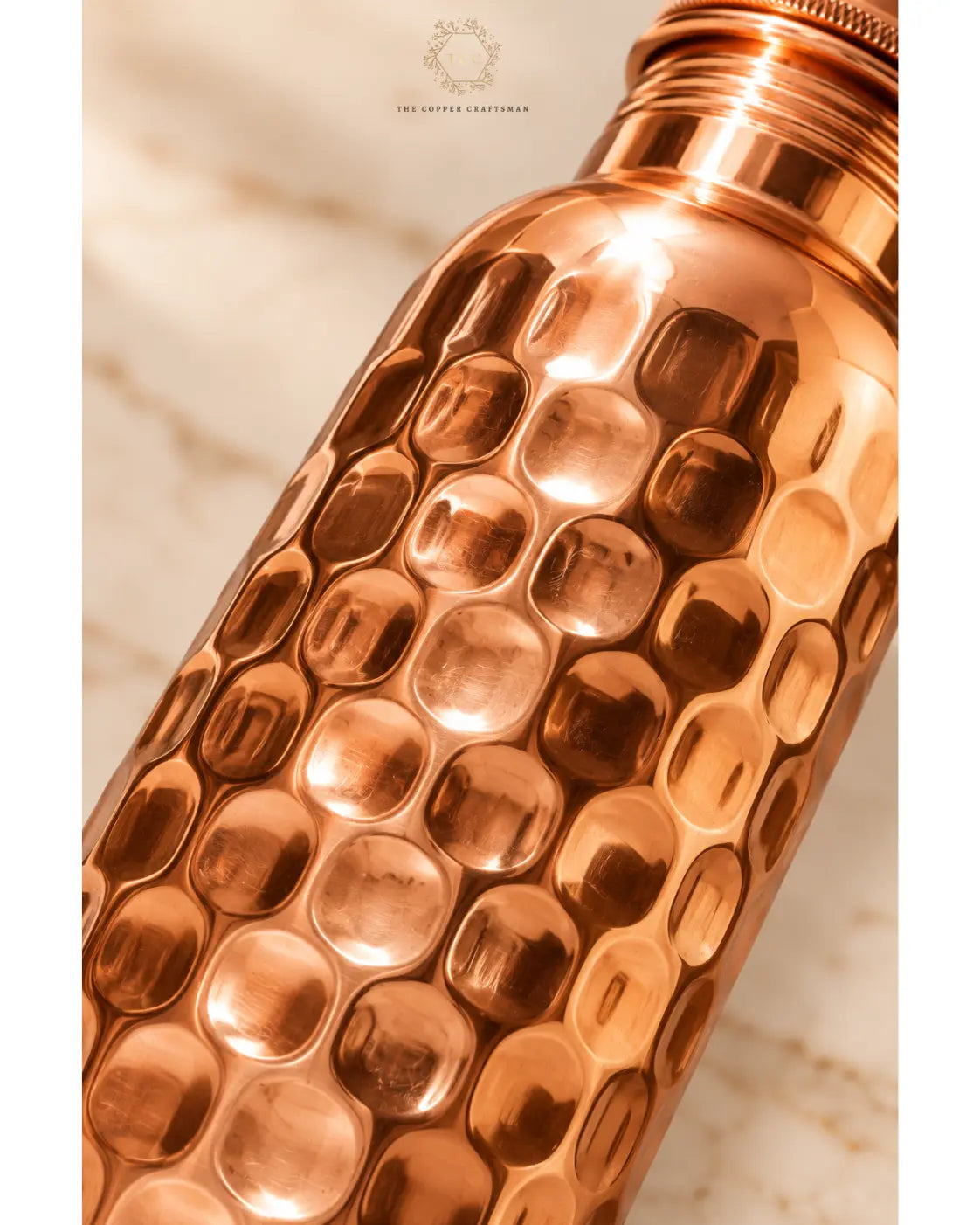 A close-up of a hammered copper tumbler with a matching metal cap, resting on a light marble surface. The brand name 'The Copper craftsman' is visible at the top.