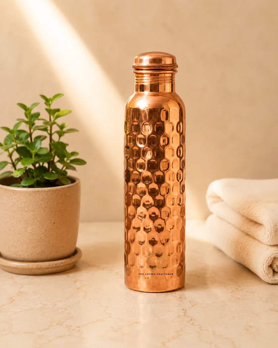A honeycomb Copper Water Bottle  with a matching lid stands on a light marble surface, flanked by a small potted green plant on the left and a folded cream towel on the right, against a warm beige wall with soft sunlight streaming in.