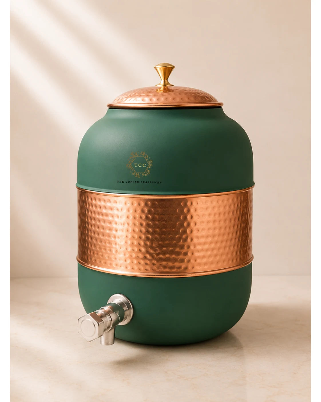 A ceramic water dispenser with a matte dark green body, a hammered copper band around the middle, a matching copper lid with a gold finial, and a silver metal spout. The brand logo 'The Copper Conservatory' is visible on the front.
