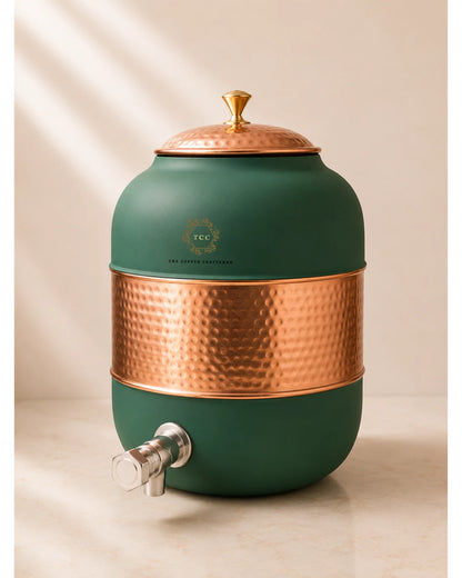 A ceramic water dispenser with a matte dark green body, a hammered copper band around the middle, a matching copper lid with a gold finial, and a silver metal spout. The brand logo 'The Copper Conservatory' is visible on the front.