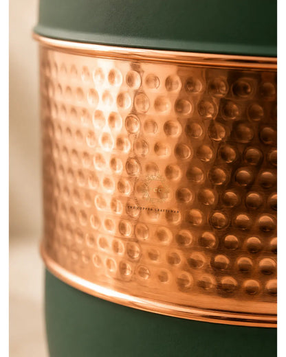 Close-up of a hammered copper band wrapped around a dark green container or vessel, with a small embossed logo in the center of the copper section.
