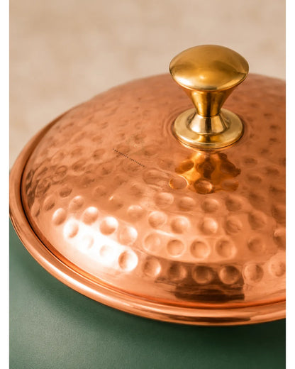 Close-up of a hammered copper lidded pot with a polished brass knob, set against a split background of beige and dark green.