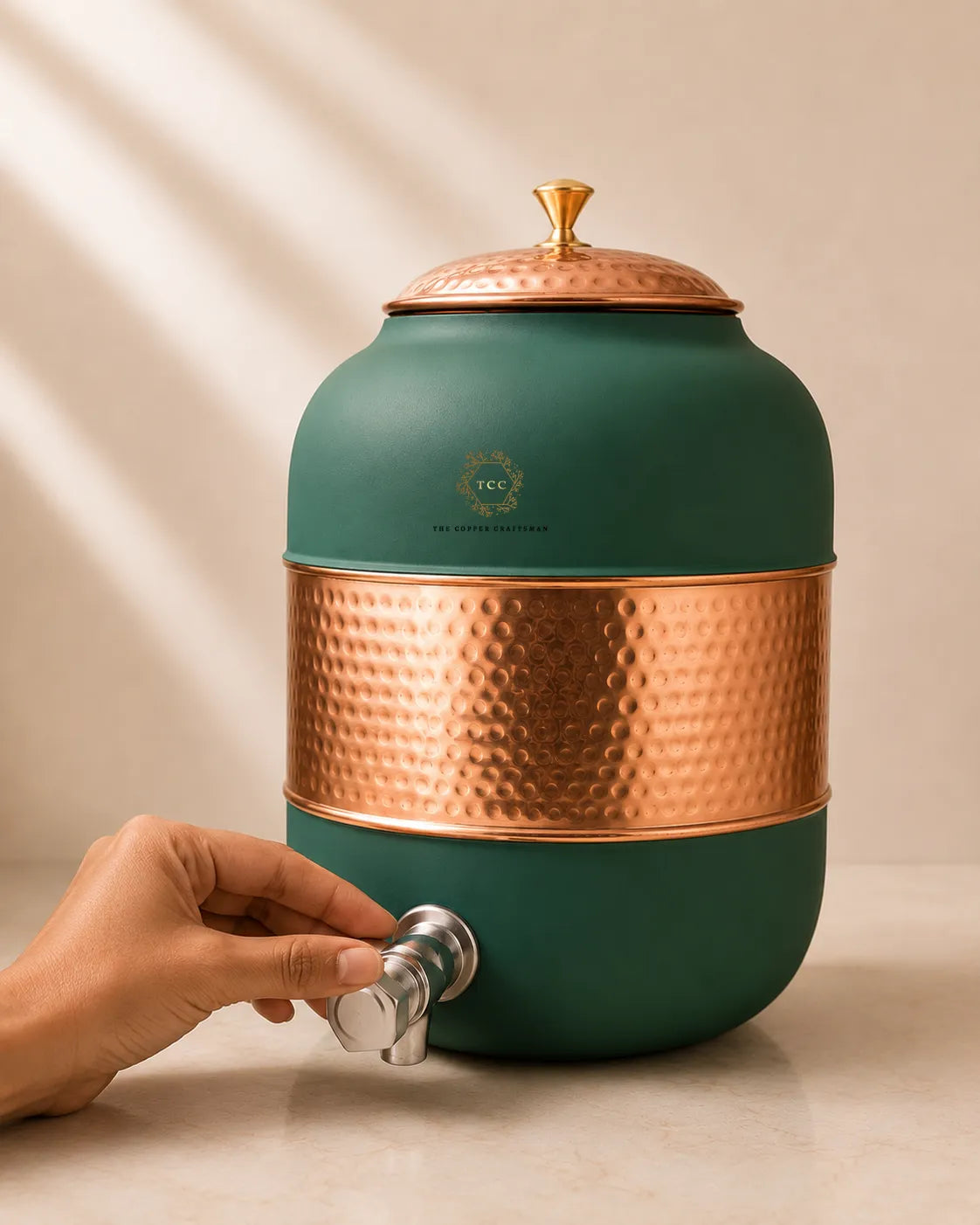 A large round green water dispenser with a hammered copper body band, a copper lid, and a silver metal spout, shown with a hand turning the spout. The brand logo 'The Tea Society' is visible on the front.