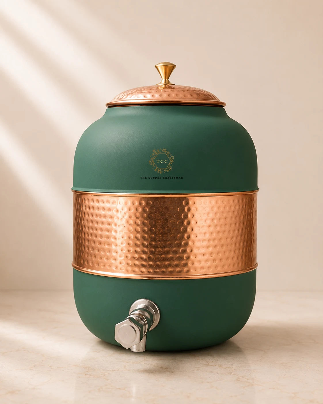 A matte green ceramic water dispenser with hammered copper accents on the lid, midsection, and spigot, shown on a light marble surface with soft diagonal shadows.