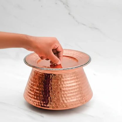 Copper cake stand with a hand placing a lid on it against a white marble background