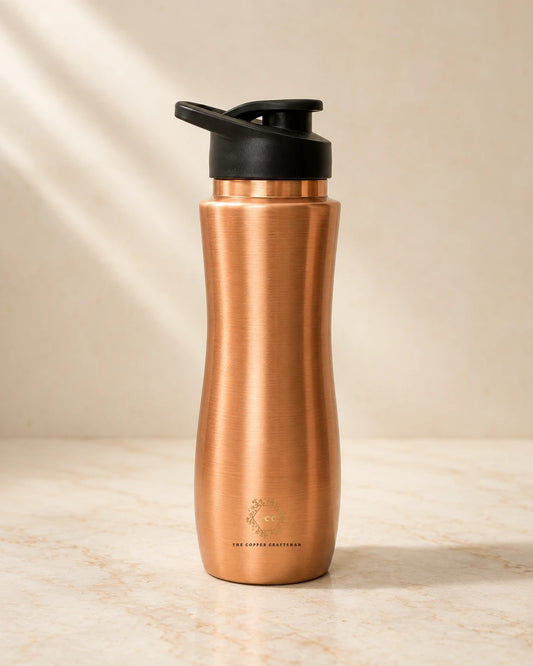 A copper water bottle with a black flip-top lid, standing on a light marble surface against a warm beige background with soft diagonal shadows.