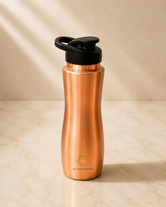 A copper water bottle with a black flip-top lid, standing on a light beige marble surface with soft diagonal shadows.