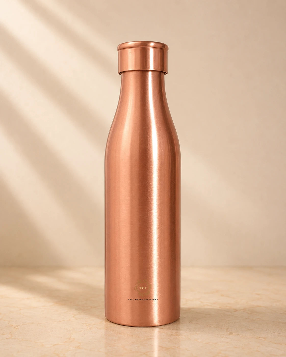 A sleek, brushed Traditional Pure Copper Water Bottle 1 L with a matching screw-on lid, standing on a light beige surface against a warm beige background with soft diagonal shadows.