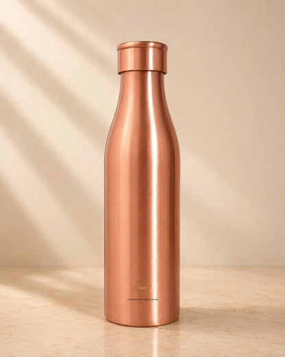 A sleek, brushed Traditional Pure Copper Water Bottle 1 L with a matching screw-on lid, standing on a light beige surface against a warm beige background with soft diagonal shadows.