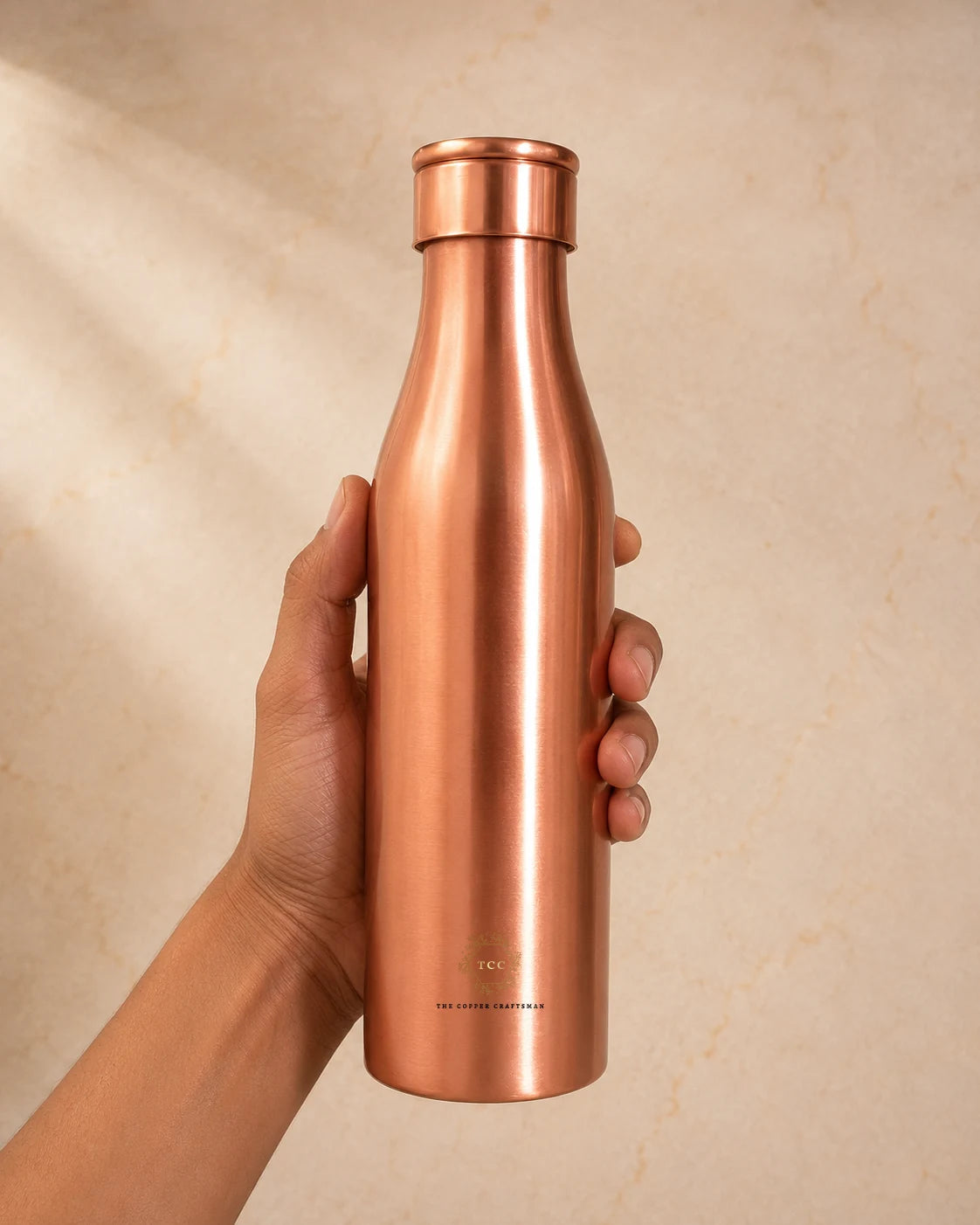 A hand holding a sleek, Traditional Pure Copper Water Bottle 1 L against a warm beige textured background.