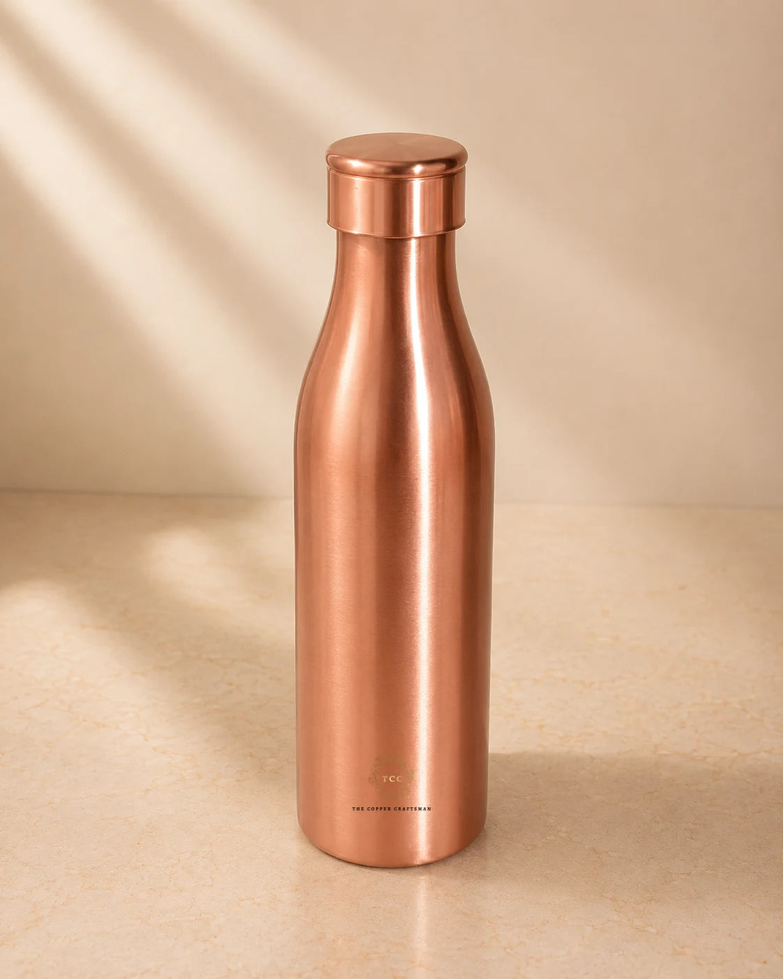 A sleek, Traditional Pure Copper Water Bottle 1 L with a matching screw-on lid, standing on a light beige surface with soft diagonal shadows.