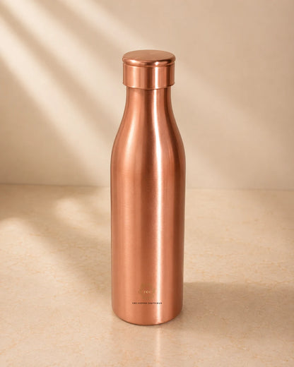 A sleek, Traditional Pure Copper Water Bottle 1 L with a matching screw-on lid, standing on a light beige surface with soft diagonal shadows.