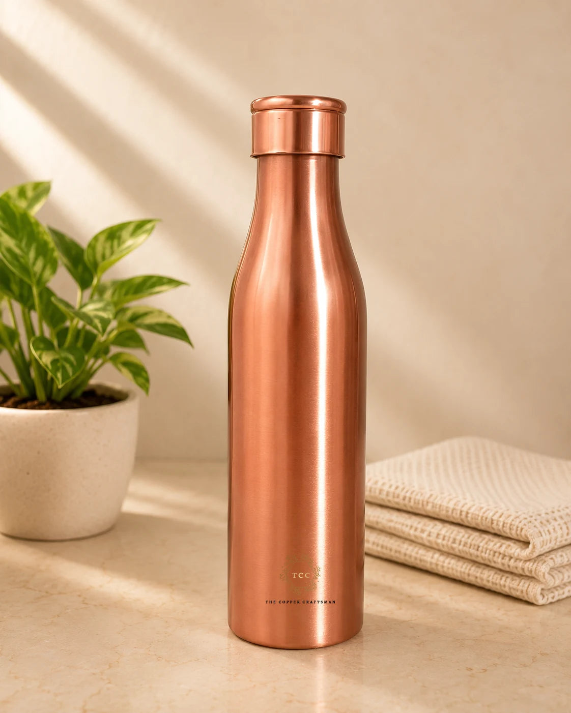 A sleek, Traditional Pure Copper Water Bottle 1 L with a matching screw-on lid, standing on a light beige surface. The background is a warm beige wall with soft diagonal shadows, flanked by a potted green plant on the left and a folded cream-colored textured towel on the right.
