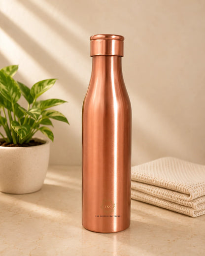 A sleek, Traditional Pure Copper Water Bottle 1 L with a matching screw-on lid, standing on a light beige surface. The background is a warm beige wall with soft diagonal shadows, flanked by a potted green plant on the left and a folded cream-colored textured towel on the right.