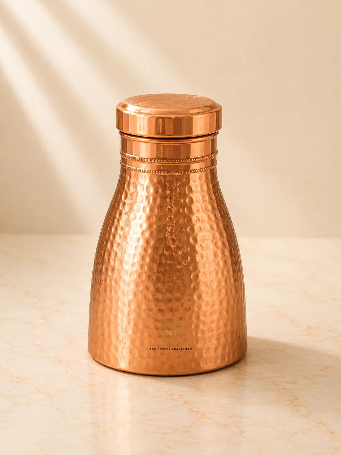 A hammered Copper Bedroom Jar 1L standing on a light marble surface against a warm beige background with soft diagonal shadows.