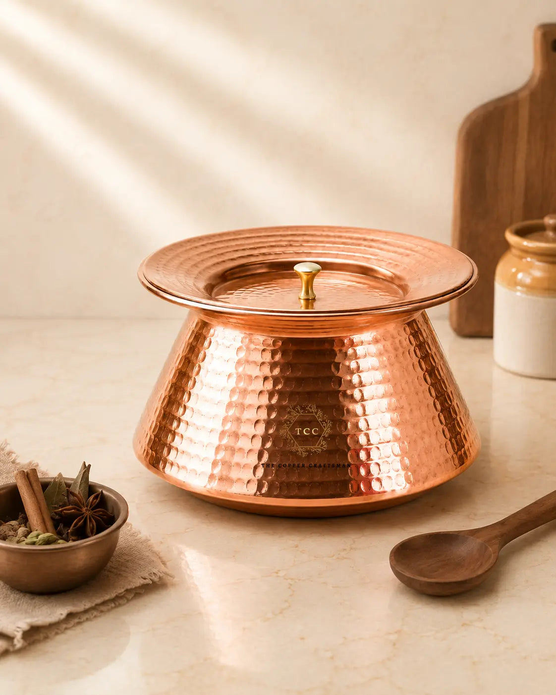 A copper-handi-with-tin-lining with a small gold-toned knob, styled on a light marble surface with a wooden spoon, spices, and a ceramic jar in the background.