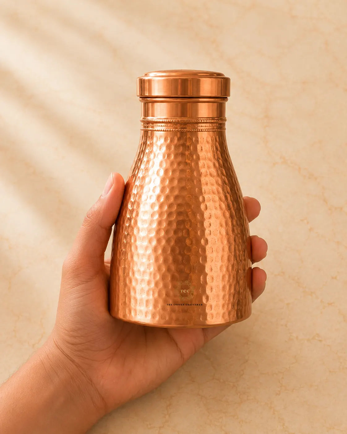 A hand holding a small Copper Bedroom Jar 1L  against a warm beige background with soft leaf shadows.