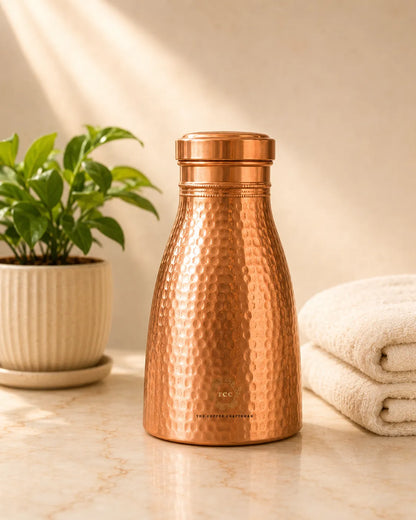 A hammered Copper Bedroom Jar 1L with a matching lid, standing on a marble surface with a potted plant on the left and folded white towels on the right.