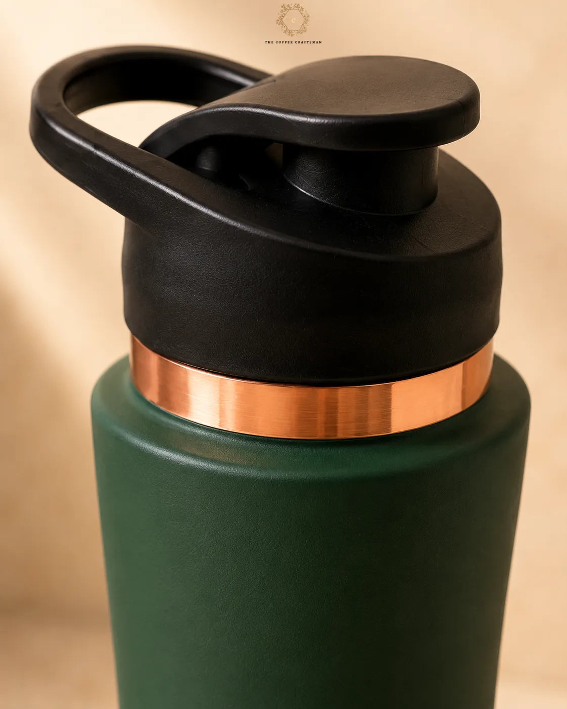 Close-up of a copper bottle  with a matte dark green body, a metallic copper band around the neck, and a black flip-top lid with a carrying loop, shown against a warm beige background.