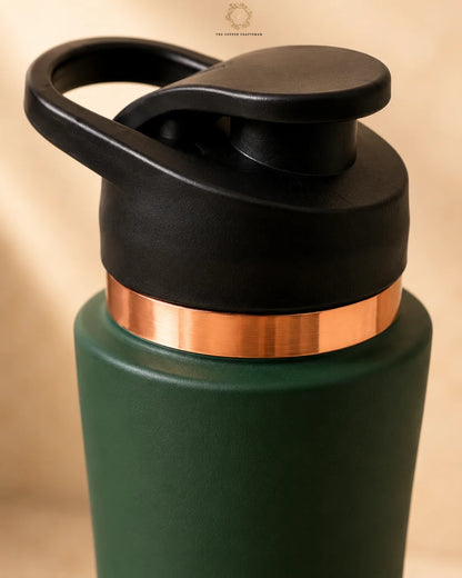 Close-up of a copper bottle  with a matte dark green body, a metallic copper band around the neck, and a black flip-top lid with a carrying loop, shown against a warm beige background.