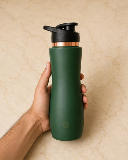A hand holding a matte dark green insulated copper bottle  with a black flip-top lid and a metallic copper collar, set against a beige stone-textured background.