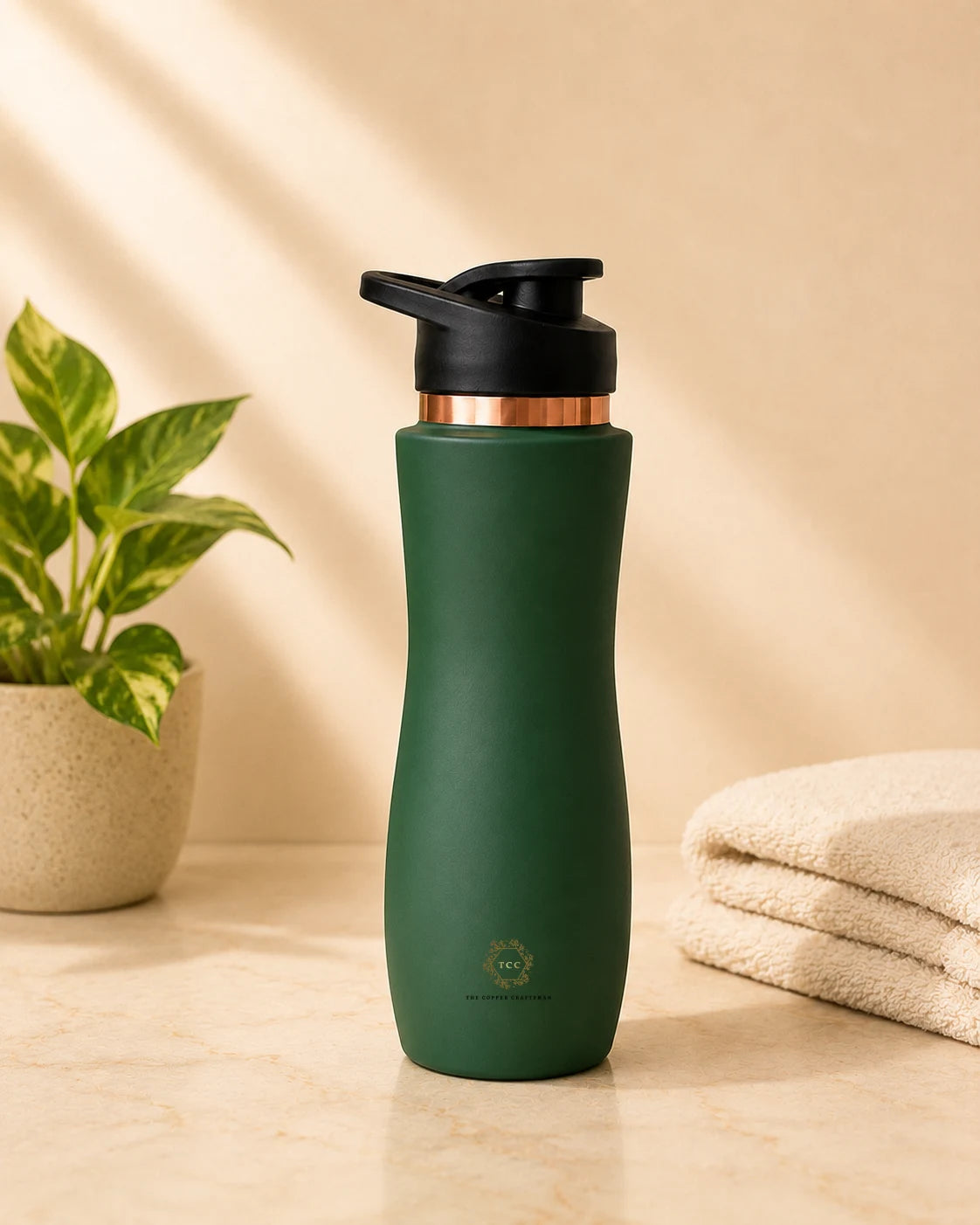 A dark green insulated copper bottle  with a black flip-top lid and a metallic rose gold collar, standing on a beige marble surface next to a potted plant and folded towels.