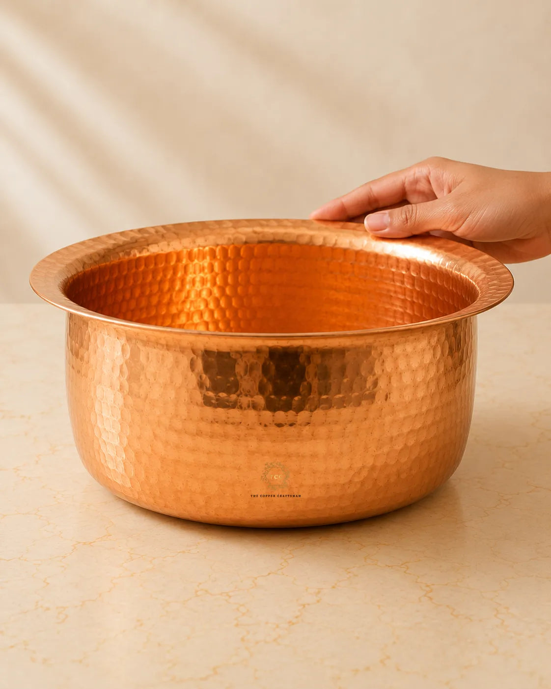 A hammered copper patila with a flared rim, shown on a light beige marble-like surface with a hand resting on the edge. The background is a soft beige fabric backdrop.