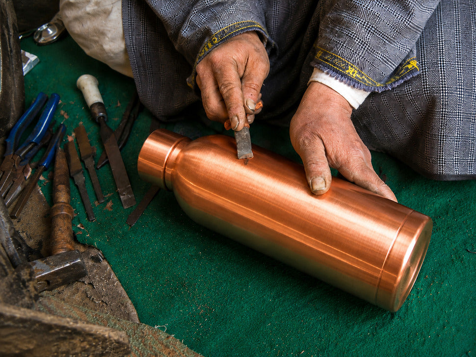 A close-up of a craftsman's hands using a chisel on a shiny copper-colored metal bottle or canister placed on a green work surface, with tools nearby.