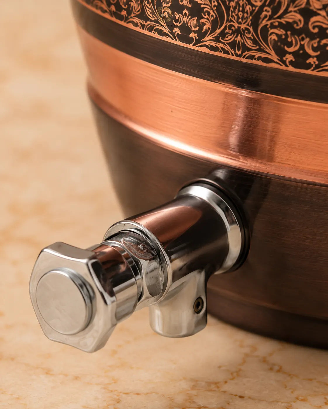 Close-up of a copper and dark brown ceramic beverage dispenser with a shiny chrome spout and hexagonal handle, set on a beige marble surface.