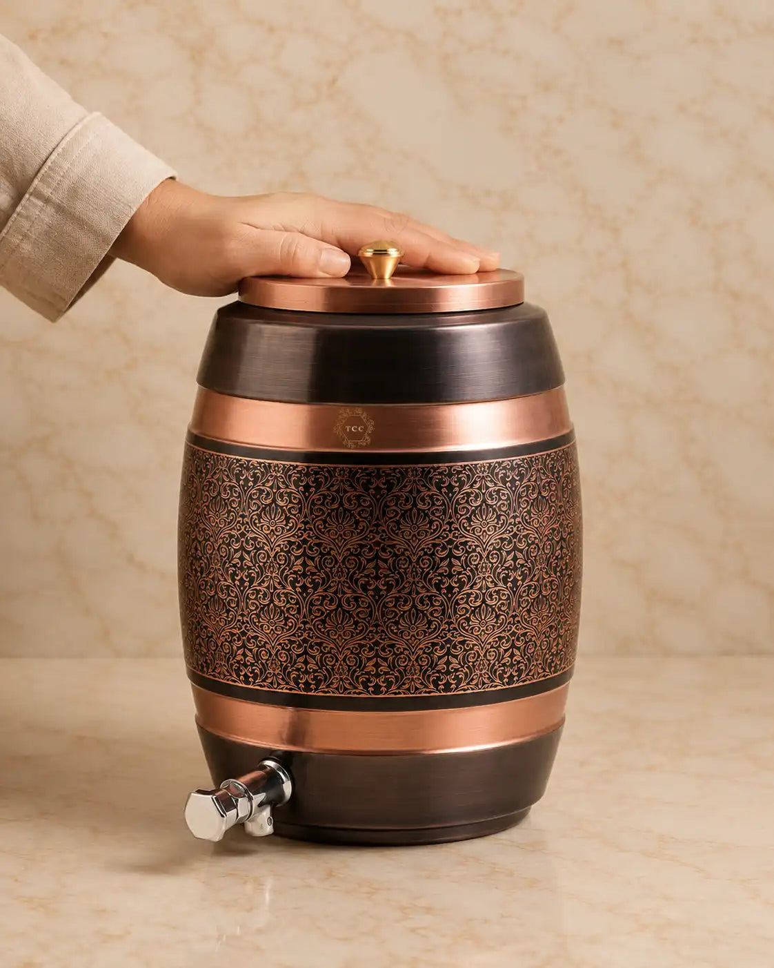 A Copper Water Dispenser shaped like a barrel, with an ornate cutout pattern on the body, a lid with a small gold knob, and a metal spout with a hex key. A hand is resting on the lid.