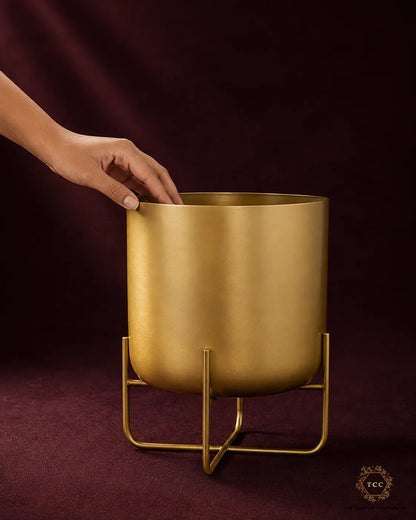 A matte gold cylindrical planter on a matching metal stand, held by a person's hand against a dark burgundy backdrop.