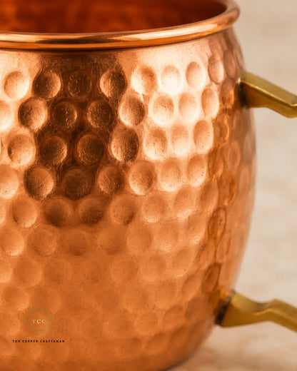 Close-up of a hammered copper Moscow Mule mug with a polished brass handle, showing the textured metallic surface and warm copper tones against a soft neutral background.
