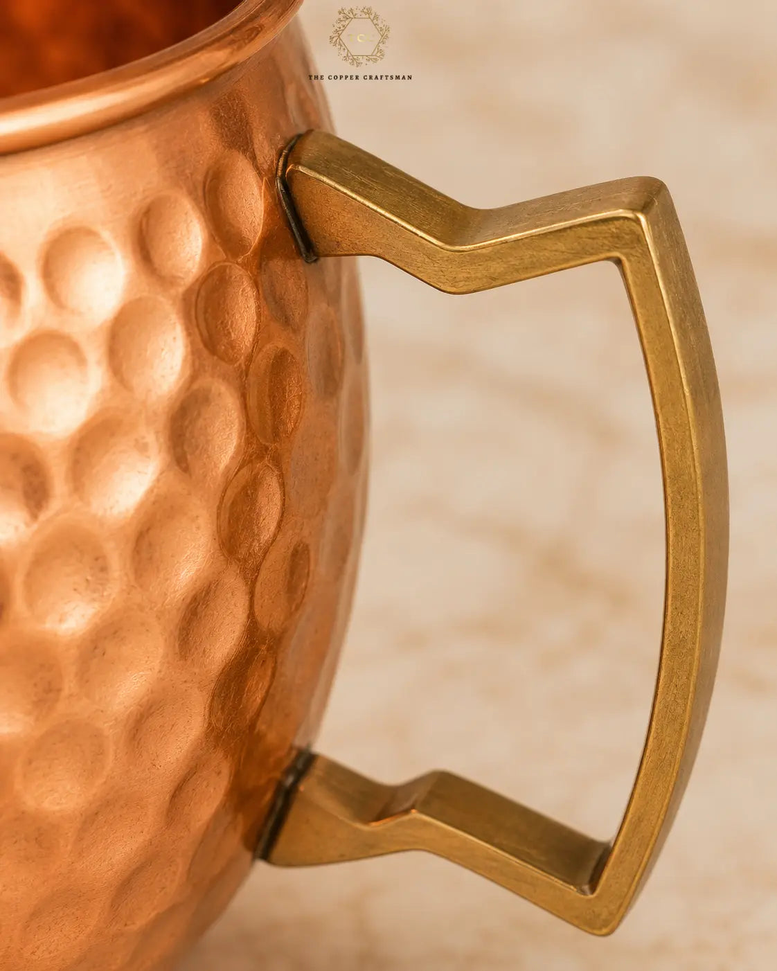 Close-up of a hammered copper mug with a geometric brass handle, set against a light beige marble background. A logo for 'The Copper Collection' is visible at the top.