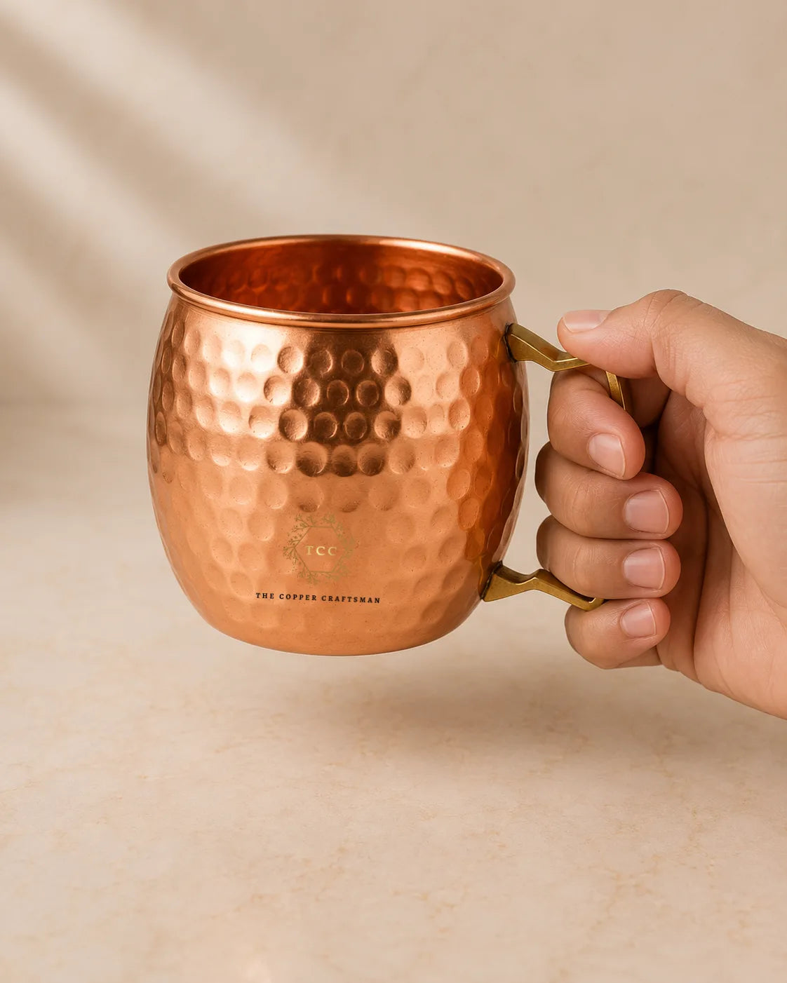 A hand holding a hammered copper Moscow Mule mug with a gold-toned handle, set against a warm beige background.