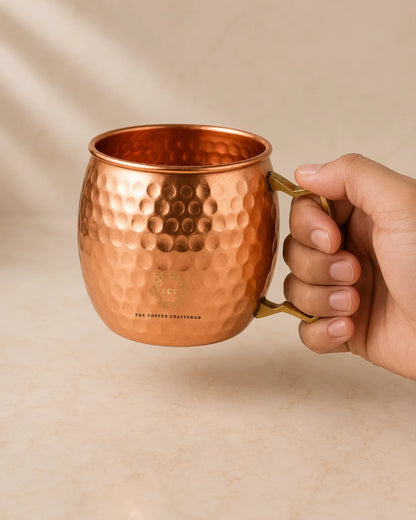 A hand holding a hammered copper Moscow Mule mug with a gold-toned handle, set against a warm beige background.