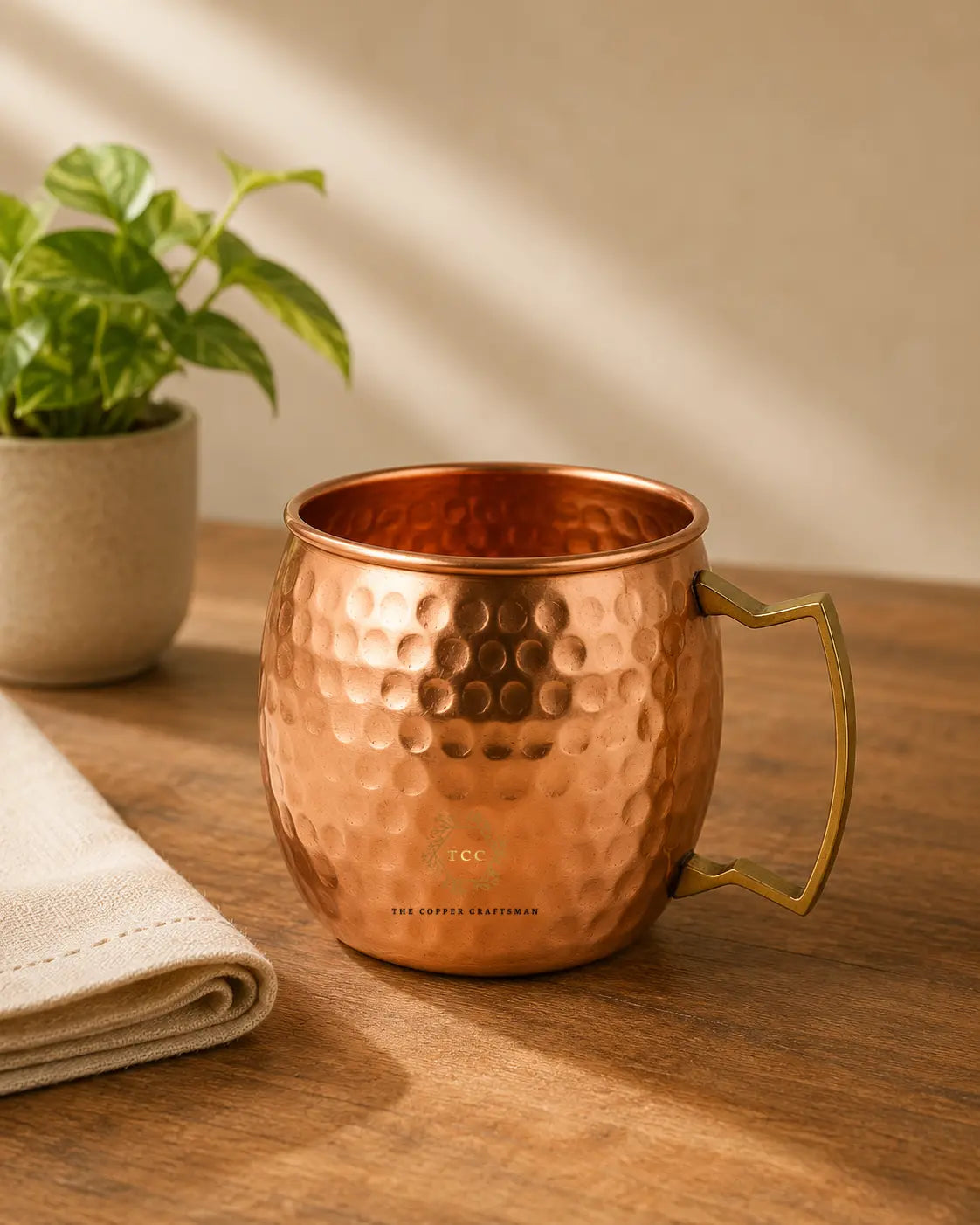A hammered copper Moscow mule mug with a brass-toned handle, set on a wooden table next to a folded napkin and a small potted plant.