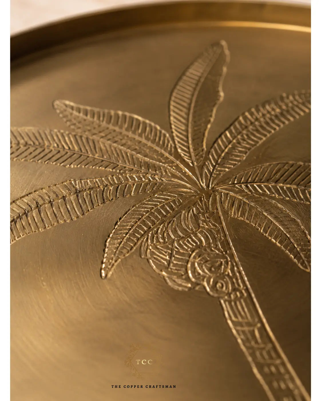 Close-up of a brass or bronze decorative tray featuring an embossed palm tree design, with a small logo and the text 'THE COPPER CRAFTSMAN' near the bottom.