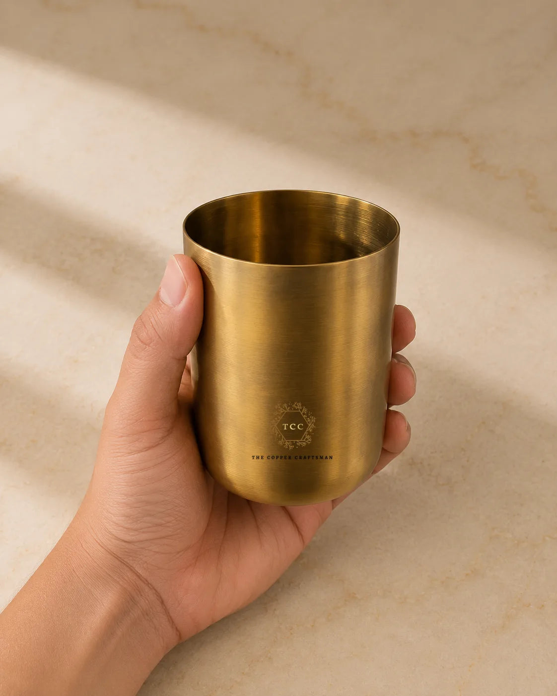 A hand holding a brushed brass tumbler cup with a visible 'TCC' logo and 'THE CRAFTED CUPBOARD' text on a beige stone surface with soft shadows.